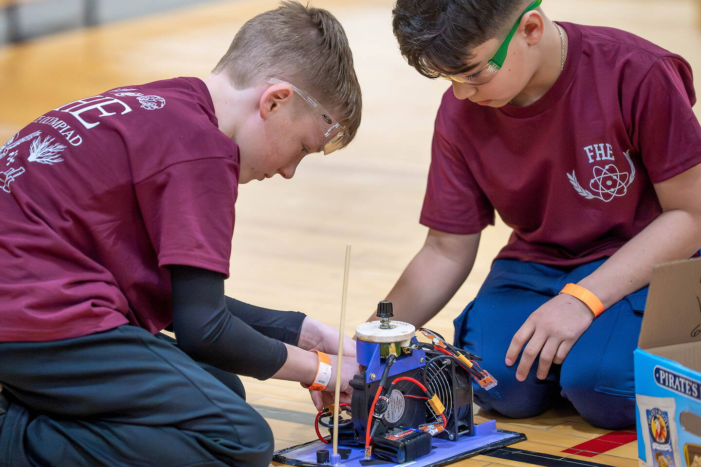 Two individuals in maroon shirts kneel on a wooden floor, focused on assembling or adjusting a small mechanical device with wires and motors.